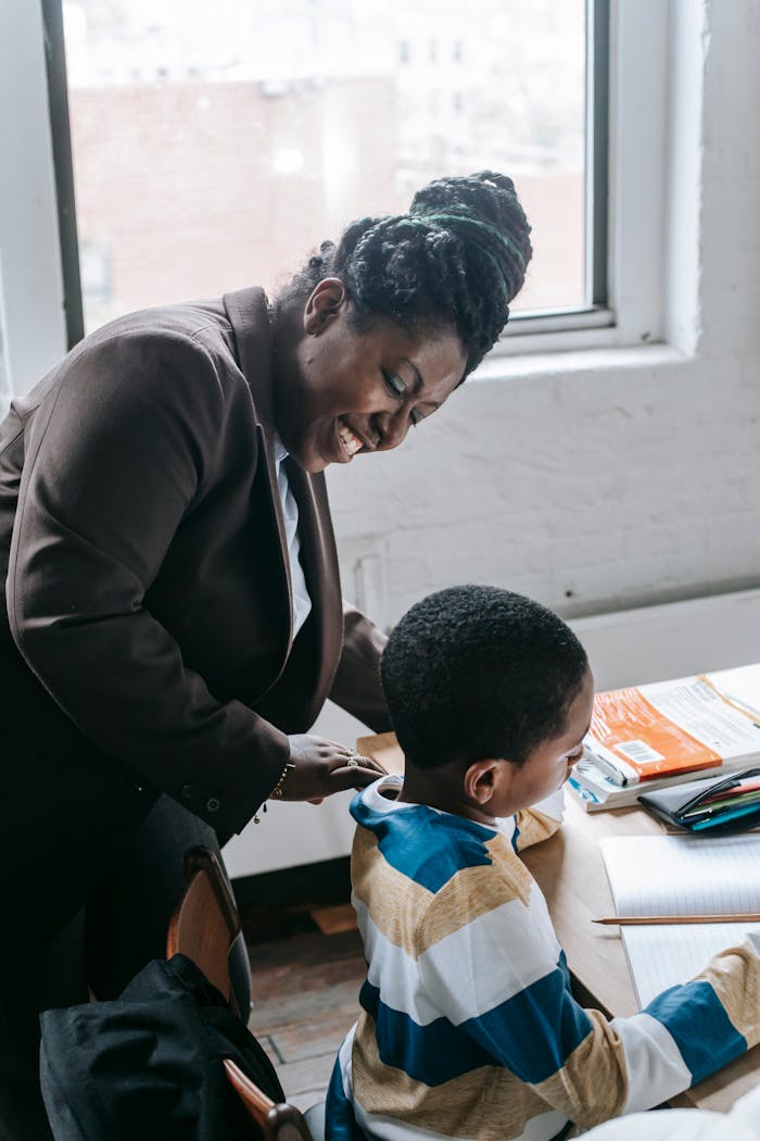 High angle of smiling adult ethnic female tutor teaching attentive little boy solving task during lesson in classroom